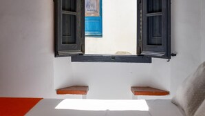 A small room with a white bed, two wooden shelves, and a window with open shutters.
