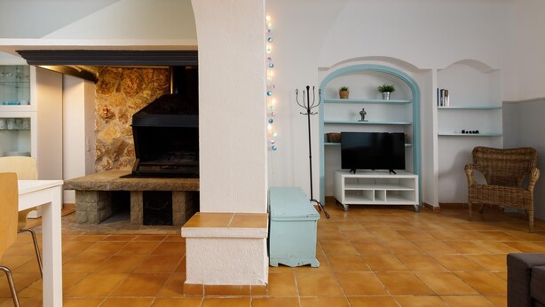 A living room with a stone fireplace, a television on a white stand, a wicker chair, and a shelf with decorative items.