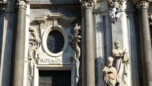 Fachada de edificio con columnas clásicas, un gran ventanal arqueado y una estatua de una figura sosteniendo un libro.
