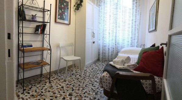 A small, neatly arranged bedroom with a bed, a white chair, a metal bookshelf, and a patterned floor.