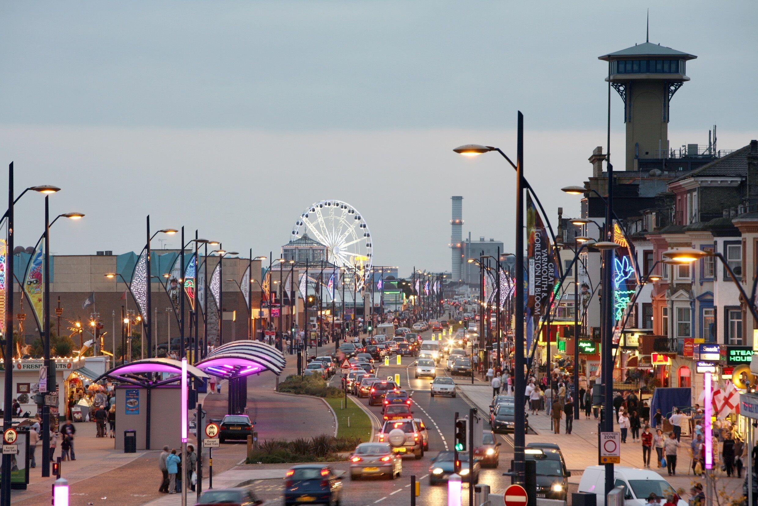 A busy urban street with a prominent Ferris wheel in the background, street lamps, and various shops and buildings.