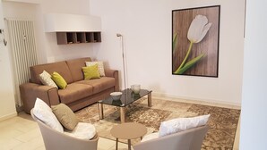 Ein Wohnzimmer mit einem braunen Sofa, zwei Sesseln, einem Glastisch und einer Stehlampe. An der Wand hängt ein Heizkörper und ein gerahmtes Bild einer Tulpe.
