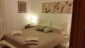 Ein ordentlich bezogenes Bett mit Kissen und gefalteten Handtüchern, ein Nachttisch mit Lampe und Wandkunst über dem Bett.