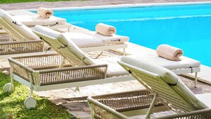 Un espace aménagé au bord de la piscine, avec des chaises longues et un grand parasol, entouré de verdure et d’un mur en pierre.
