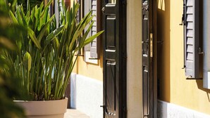 Un bâtiment à la façade jaune, aux volets noirs, avec un pot de fleurs placé au premier plan.