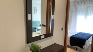 Quarto de hotel com um espelho grande, uma mesa pequena com uma planta, uma cama com colcha azul-escuro e uma janela com cortinas finas.