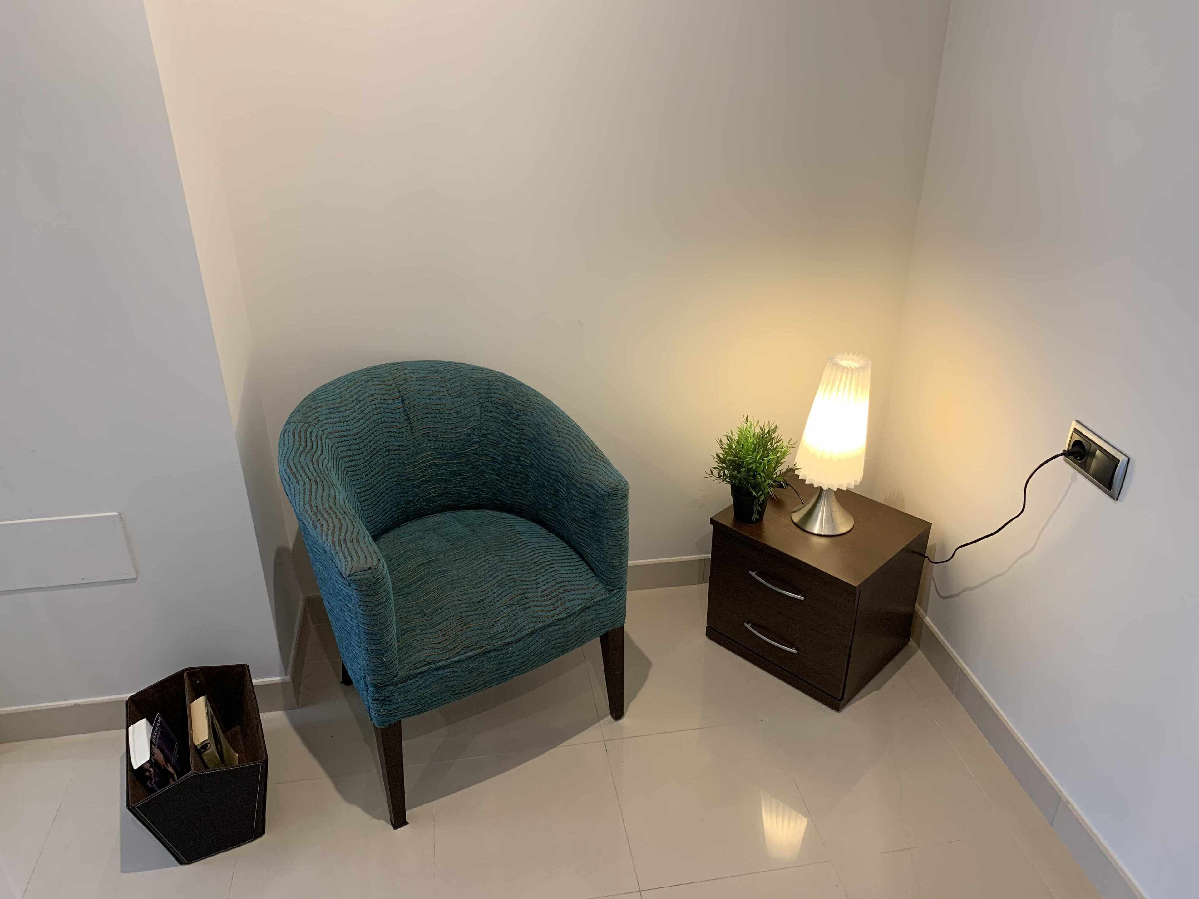 A teal armchair, a bedside table with a lamp, and a small black storage box in a room with white walls and tiled flooring.