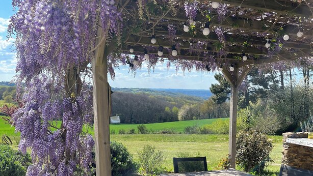 Una pérgola cubierta de hiedra morada, una mesa de madera con sillas y vistas a un campo verde y colinas al fondo.