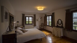 Ein Schlafzimmer mit einem Bett, einem Sessel, einem Tisch mit Spiegel und zwei Fenstern mit Vorhängen.