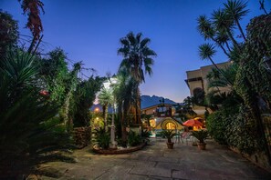 A courtyard with palm trees, a stone pathway, and a building with a yellow facade and arched entrance. - Villa Amalia – Historic Jewel of Amalfi Coast,  with Pool, Sauna & Tropical Park (Cava dei Tirreni)