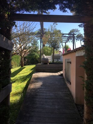 A wooden pathway leading to a covered area with a pergola overhead, surrounded by greenery and a building with a tiled roof.