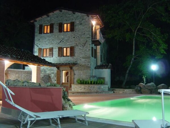 Un edificio de piedra con piscina, un área cubierta con un banco y una entrada bien iluminada.