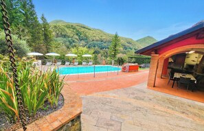 Zona de piscina con sillones de descanso y sombrillas, rodeada por un sendero de ladrillo que conduce a un edificio con techo rojo.
