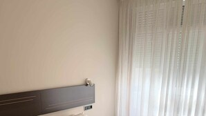 A neatly made bed with white linens and pillows, a headboard, and a window with sheer curtains.