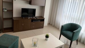 A modern living room with a flat-screen TV, wooden entertainment unit, a green armchair, and a small table with a candle and plant.