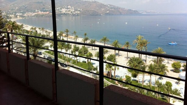 Blick aufs Meer von einem Balkon mit Palmen, Strand und einer in der Ferne liegenden Stadt.