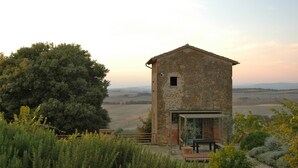 Un bâtiment rustique en pierre, doté d’un petit porche et d’un banc, entouré de verdure et offrant une vue sur une vallée.