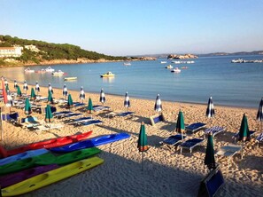 A beach with rows of sun loungers and umbrellas, a row of colorful kayaks, and boats on the water.