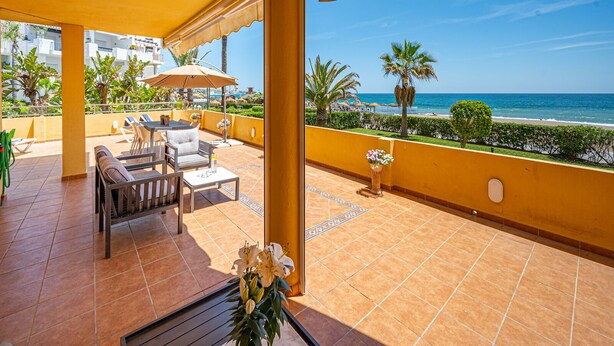 A covered terrace with outdoor seating, a view of the beach, and a pool in the background.