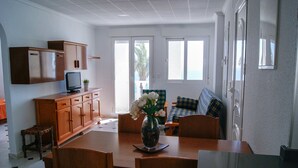 A dining area with a wooden table, chairs, a vase with flowers, and a television on a cabinet. - Apartamentos Riviera Beach (Pilar de la Horadada)