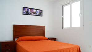 A bedroom with a wooden bed, an orange bedspread, a nightstand, a drawer, and a window with blinds. - Apartamentos Riviera Beach (Pilar de la Horadada)