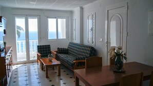 A living room with a sofa, a wooden table, and a dining table. There is a door and a window with a view of the sea. - Apartamentos Riviera Beach (Pilar de la Horadada)