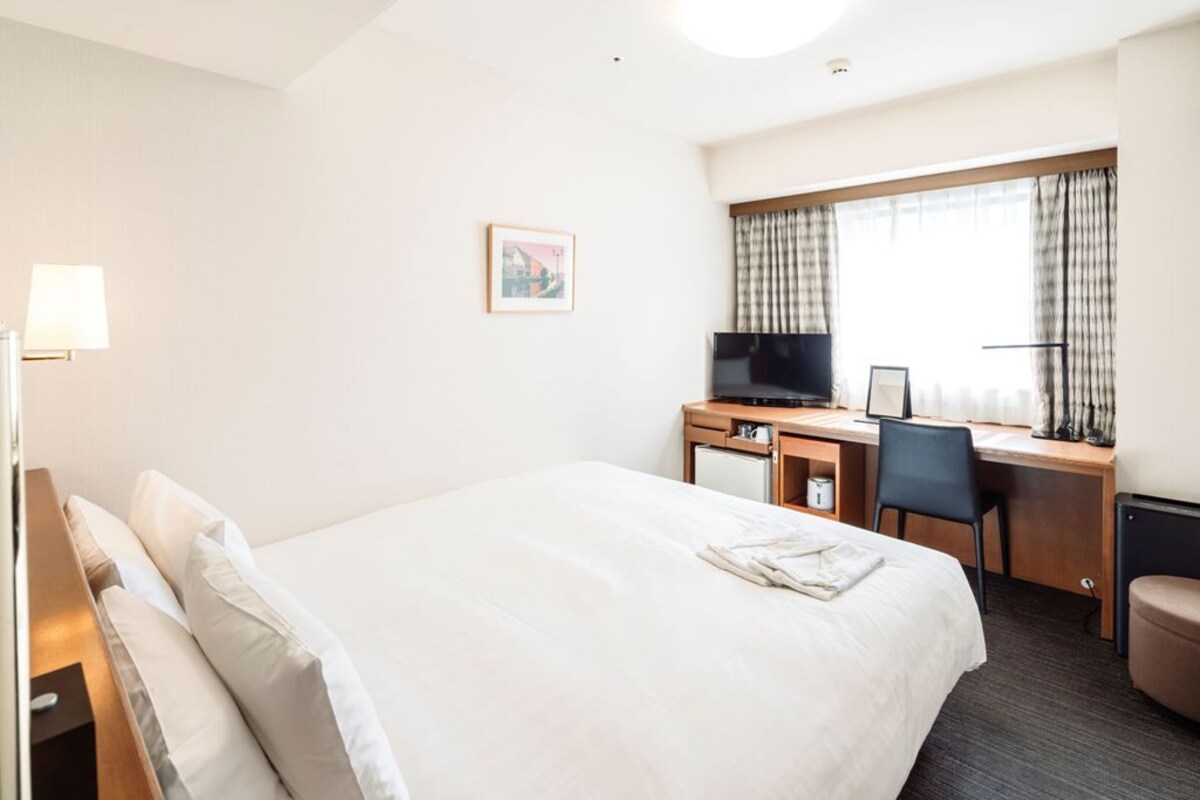Ein Hotelzimmer mit einem großen Bett, einem Schreibtisch mit Computer, einem Fernseher und einem Fenster mit Vorhängen.
