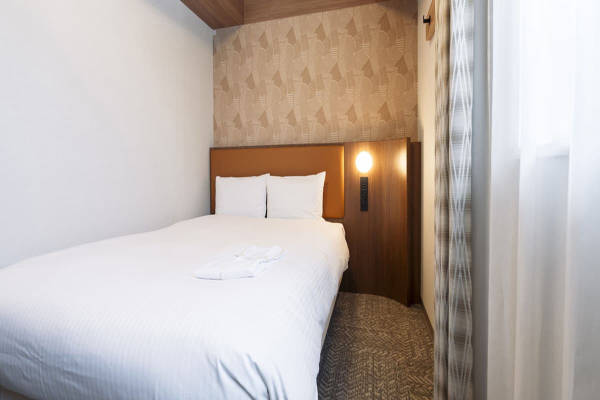 A neatly made bed with white linens and a folded towel, a wooden headboard, a patterned wall, and a wall-mounted light fixture.