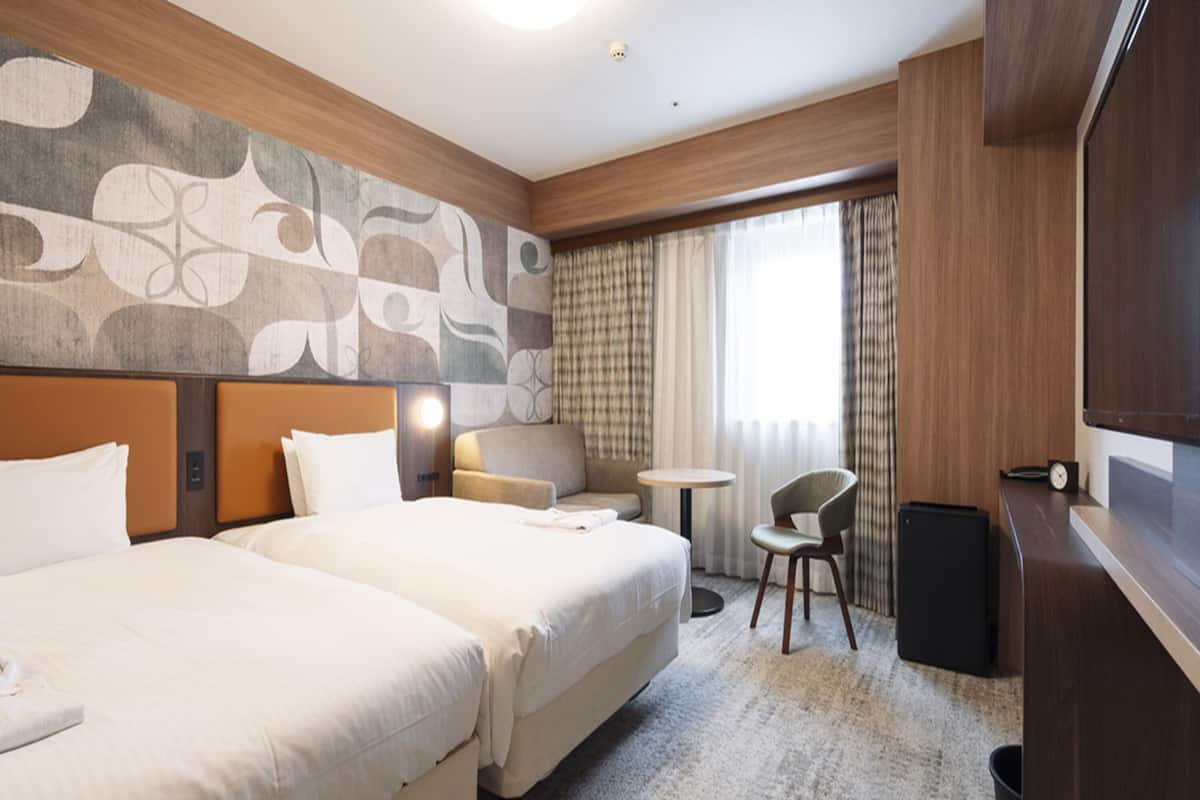 A hotel room with two beds, a sofa, a small table, a chair, a TV, and a trash bin.