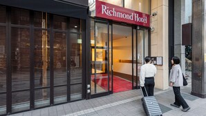 A hotel entrance with a red sign, glass doors, a red carpet, and a person pulling a suitcase.