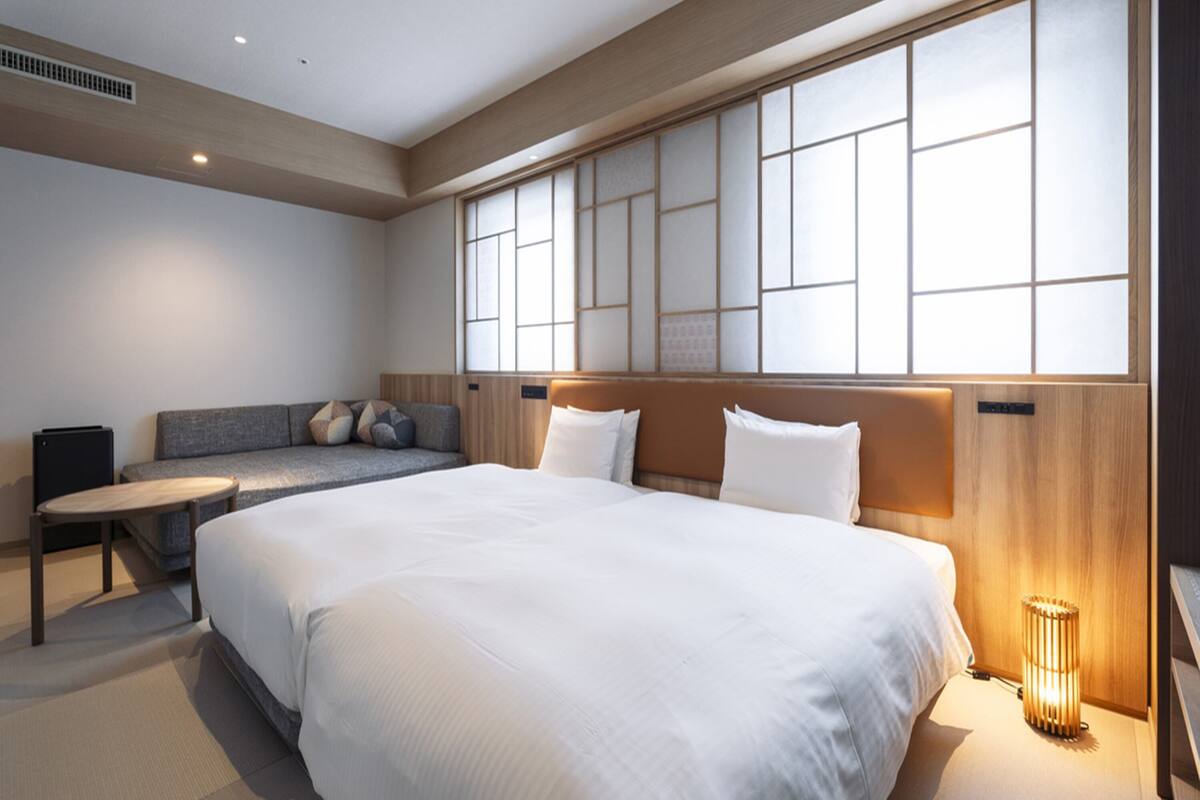 A modern hotel room with a large bed, a sofa, a small table, and a large window with a view.