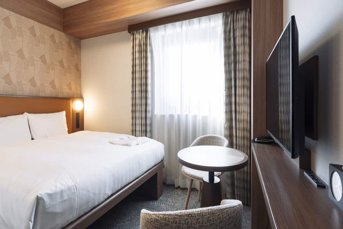 A hotel room with a bed, a small round table, a chair, a TV, and a window with curtains.