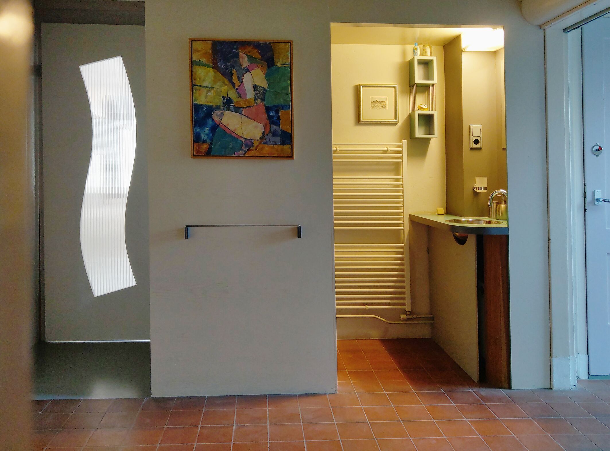 A hallway with a white door, a painting, a bathroom with a sink and mirror, and a tiled floor.