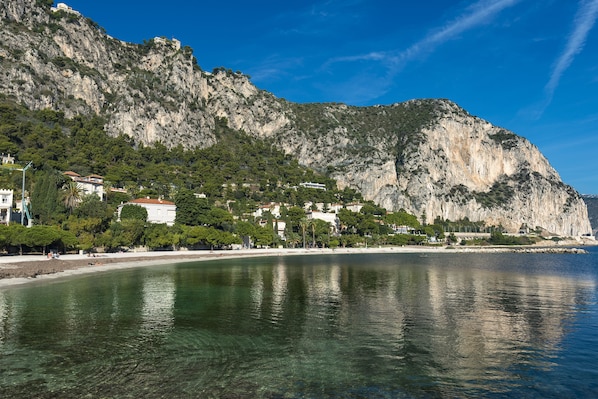 Uma vila costeira com edifícios ao longo da praia, vista clara do mar e um penhasco imponente.