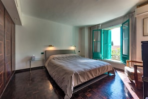 Quarto com cama, mesa de cabeceira, janela com persianas verdes e vista para o exterior.