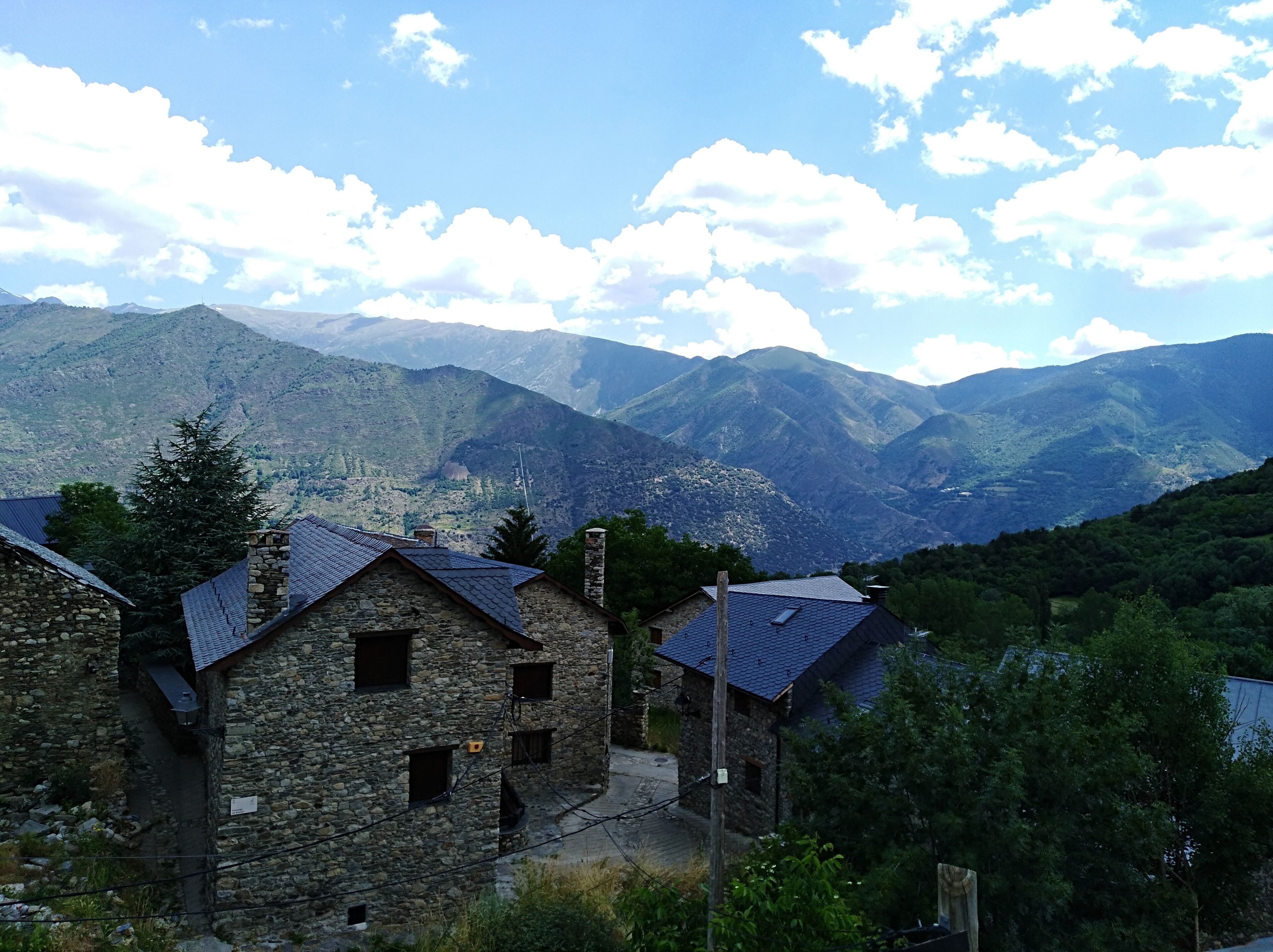 Ein Steinhäuschen mit geneigtem Dach, umgeben von üppigem Grün und Bergen im Hintergrund.