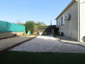 Un jardin à l’arrière d’une maison, avec un espace recouvert de gravier, une balançoire, une pelouse verdoyante et une maison dotée d’un garage attenant.