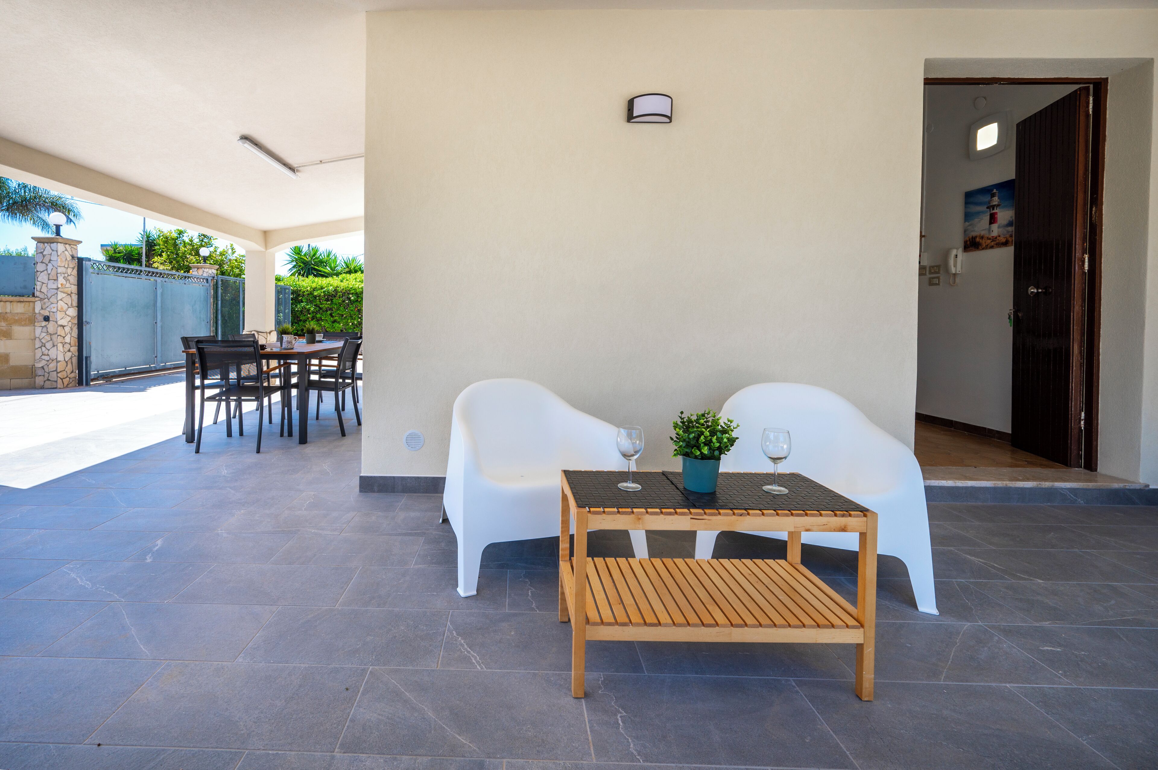 A covered outdoor seating area with a table and chairs, a potted plant, and a wine glass.