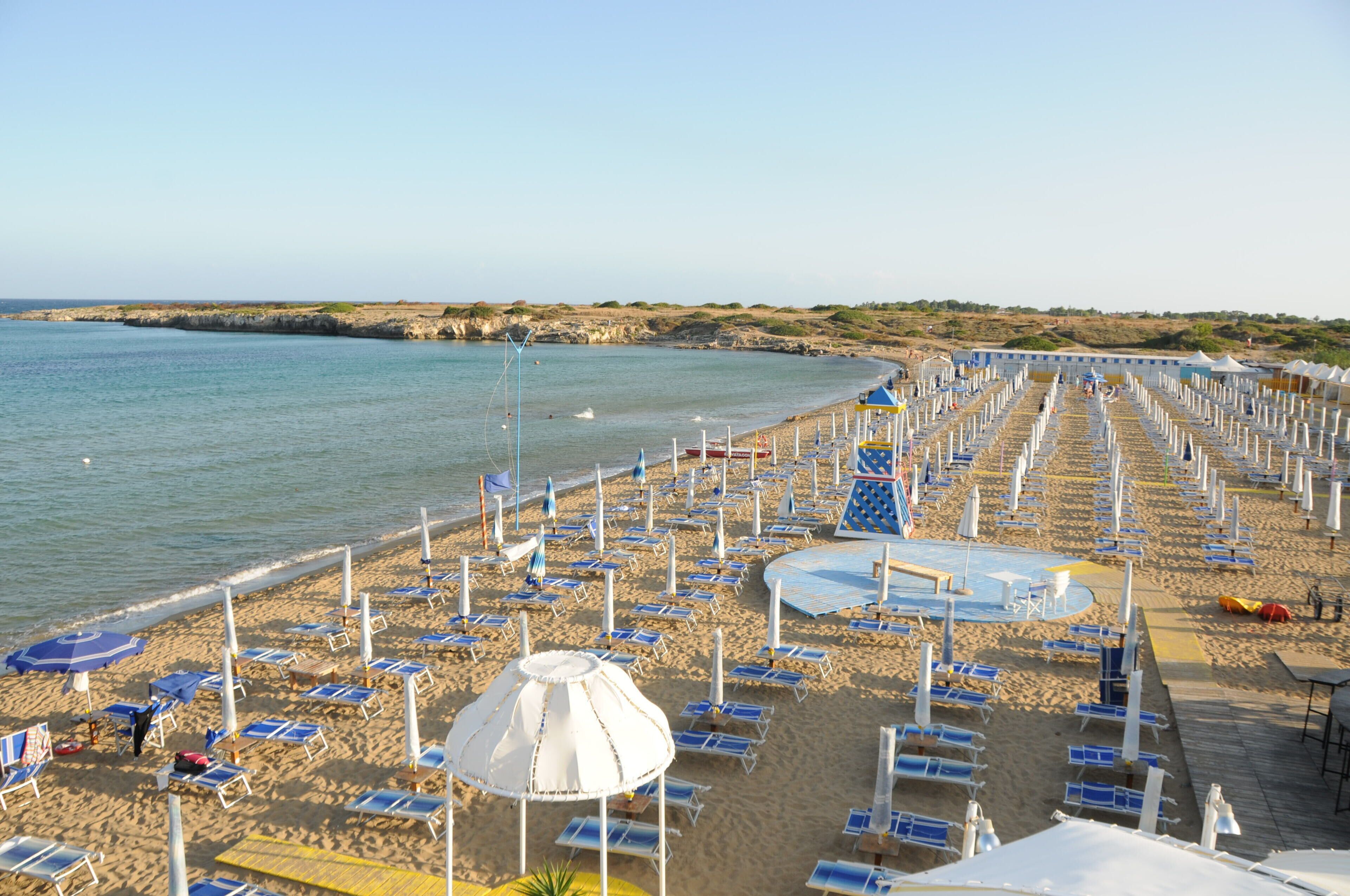 Uma praia com fileiras de espreguiçadeiras e guarda-sóis, uma piscina e um trecho de costa rochosa.