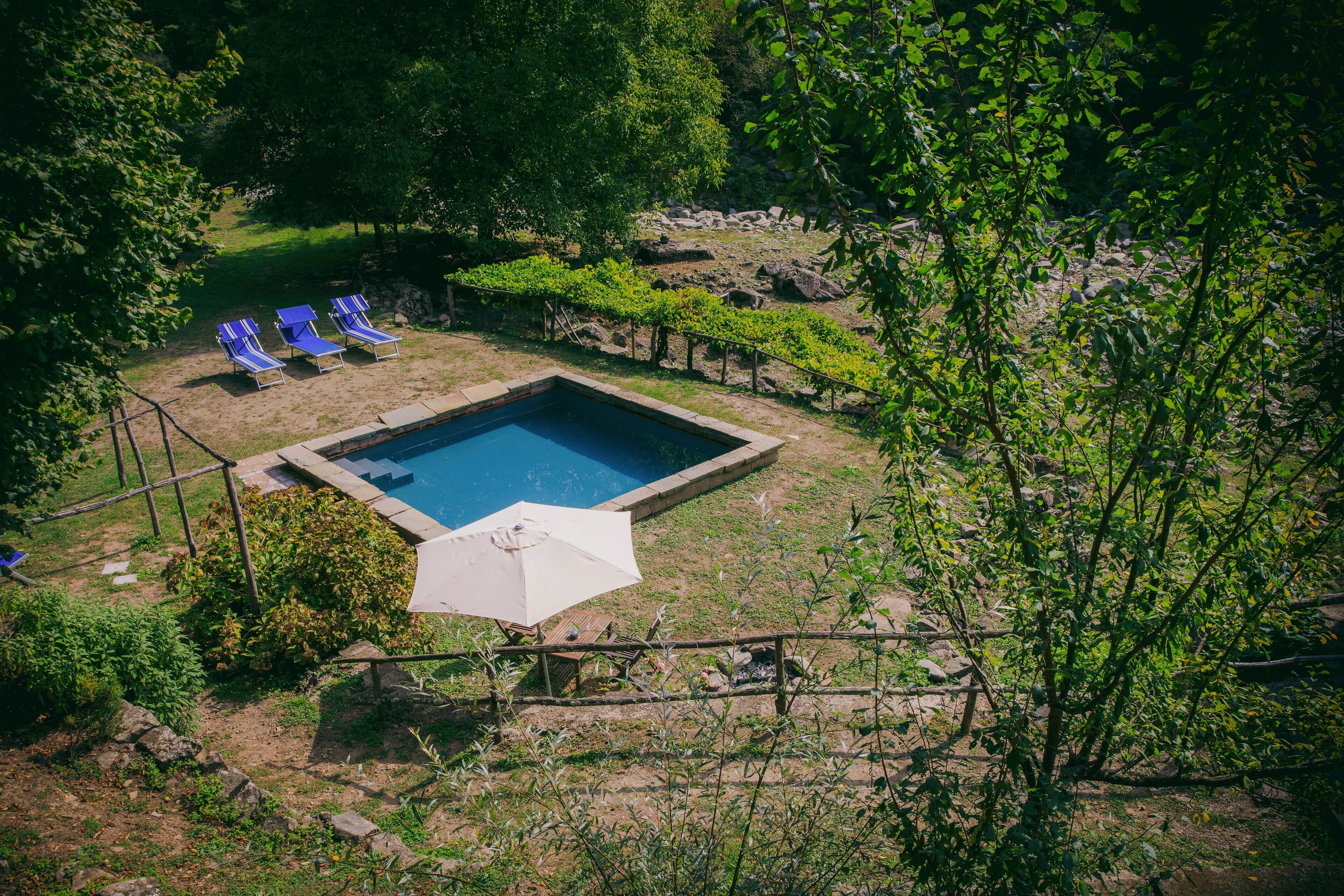 A rectangular swimming pool surrounded by a wooden fence, with three blue lounge chairs and a white umbrella.