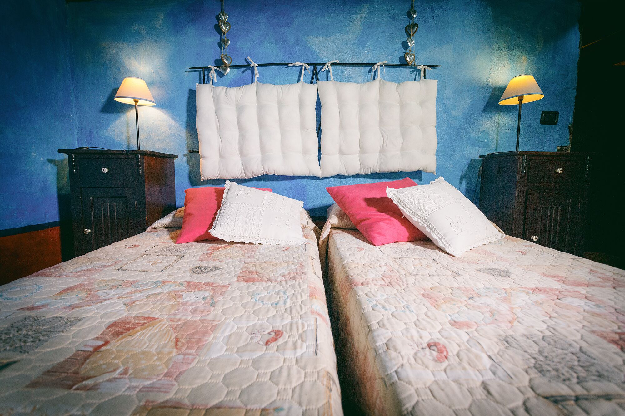 A room with two beds, each with a quilted bedspread and pillows, a blue wall, and two bedside lamps.