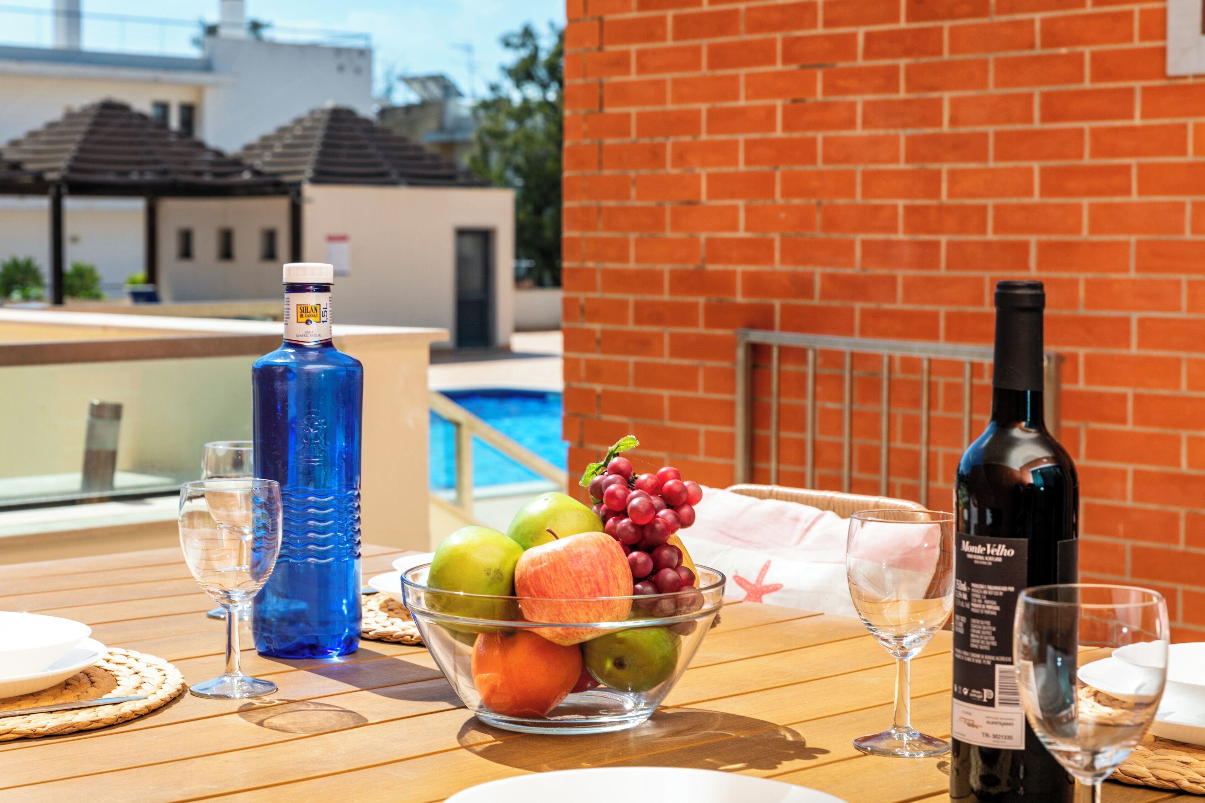 A table with a bowl of fruit, a bottle of water, and a bottle of wine, set against a brick wall with a pool in the background.