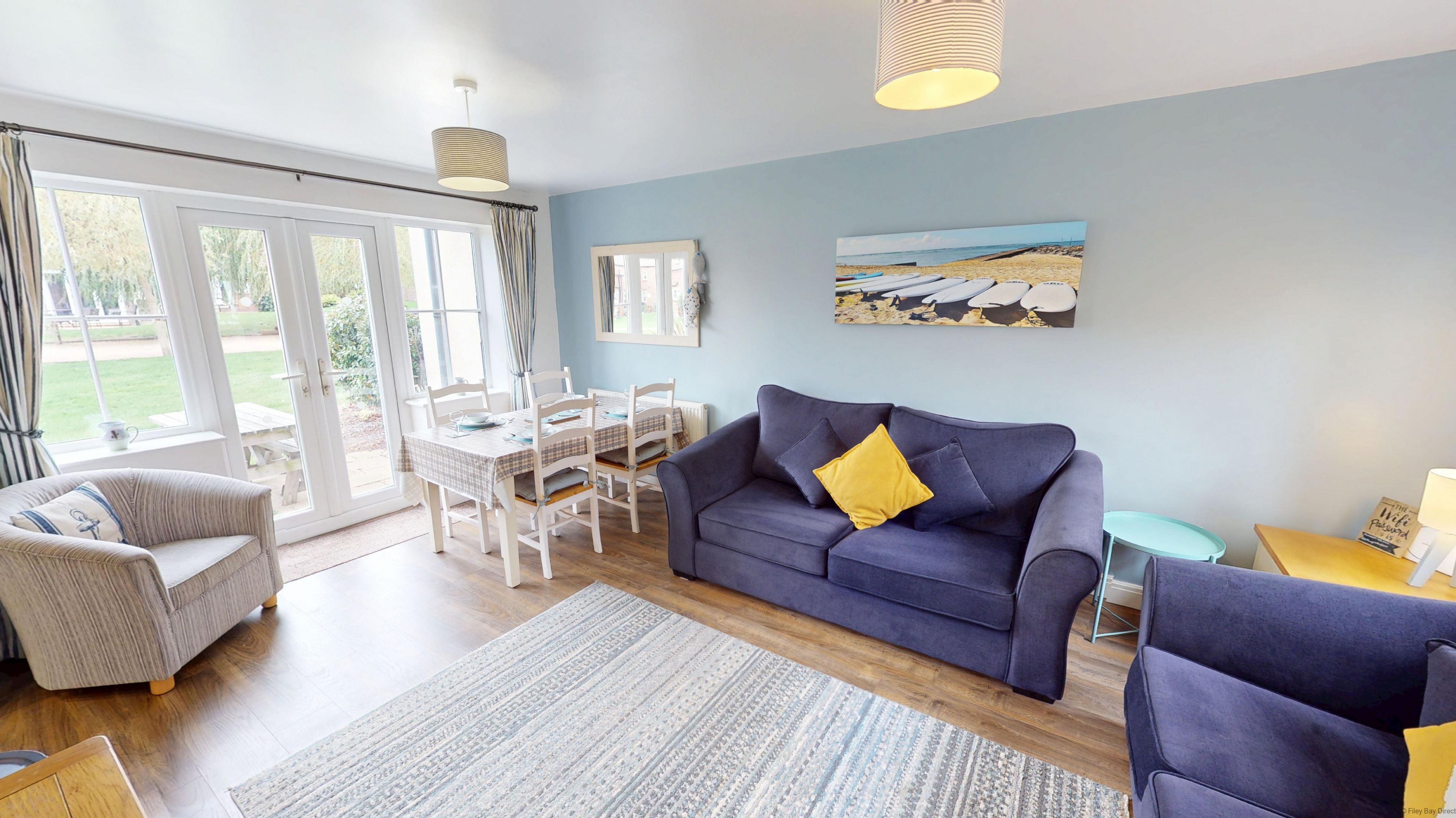 Un salon avec un canapé bleu, un fauteuil blanc, une table à manger avec des chaises et une grande fenêtre avec des rideaux.
