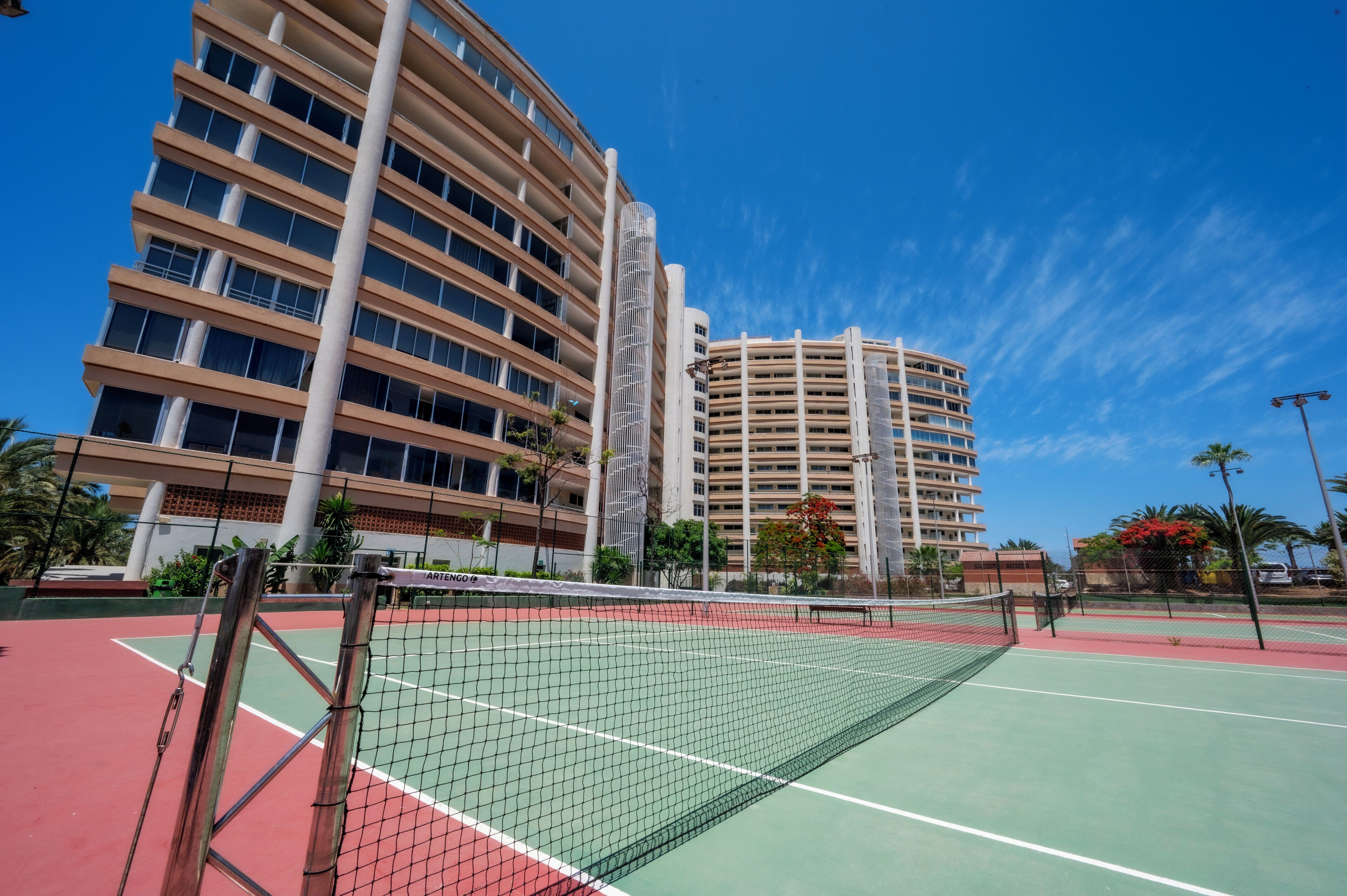A tennis court with a net, surrounded by buildings and palm trees.