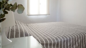 Ein schlichtes, helles Zimmer mit einem Bett, einem Nachttisch und einem Fenster mit Jalousien.