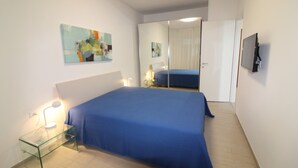 Un dormitorio con una cama grande, una mesita de noche con lámpara, un espejo y un televisor montado en la pared.