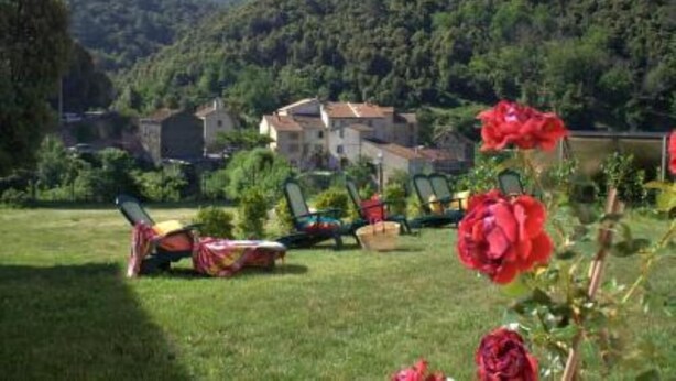 A rural setting with a large house surrounded by lush greenery and hills. There are several chairs and a table set up in the foreground, suggesting a garden or outdoor seating area.