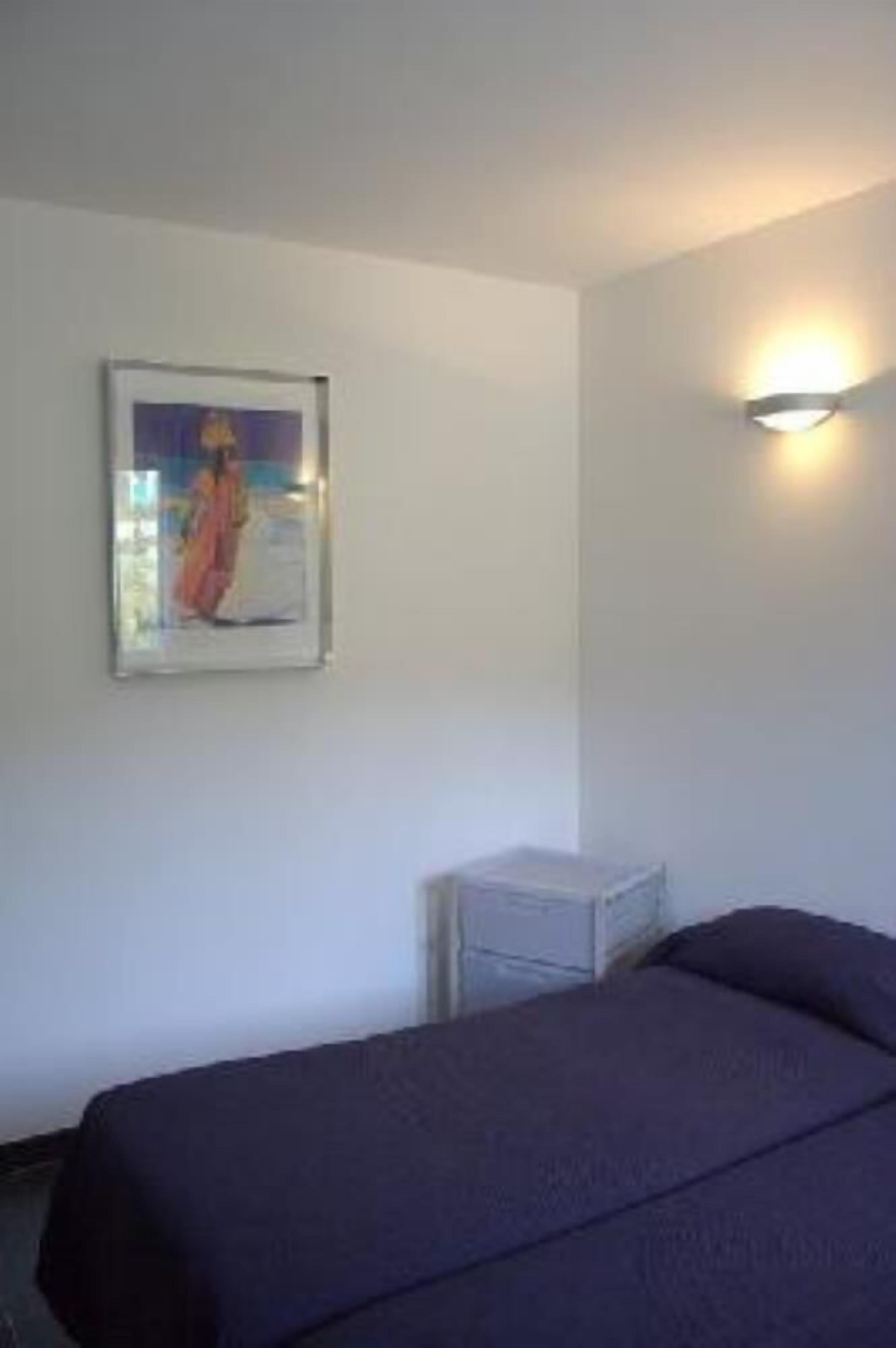 A single bed with a dark cover in a room with white walls. There is a framed picture on the wall and a small table with a lamp.