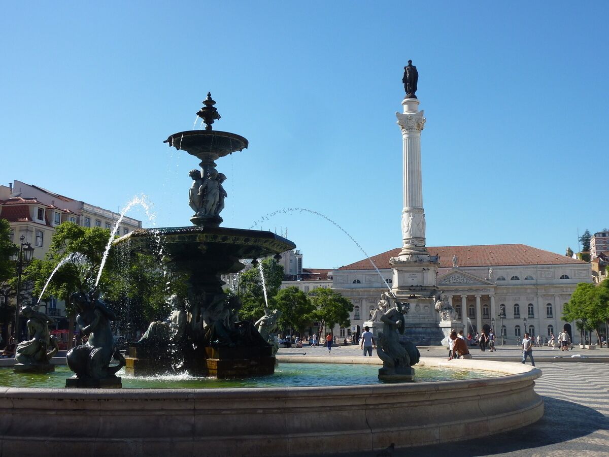 Une place publique avec une fontaine centrale, une statue sur un haut piédestal et des bâtiments qui l’entourent.