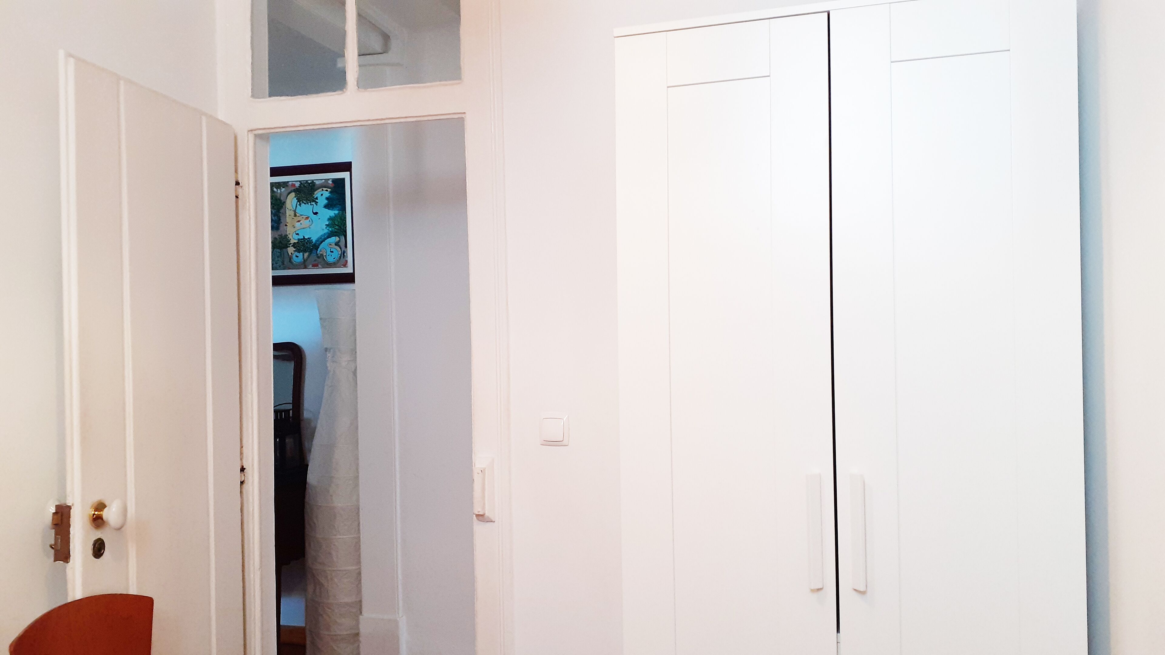 Eine weiße Tür mit kleinem Fenster darüber, die zu einem Zimmer mit Bett und Stuhl führt.